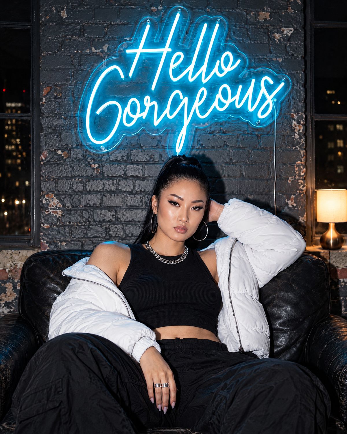 Woman in a white puffer jacket sitting under an ice blue Hello Gorgeous LED neon sign